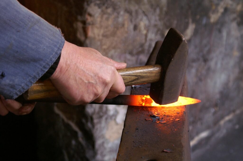A blacksmith shaping glowing metal using a hammer on an anvil inside a forge.